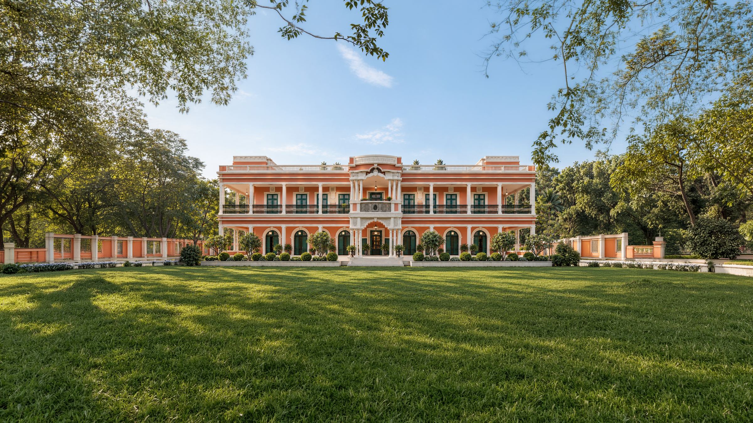 The front of the school across its lawn: two storeys in peach and white, a colonnaded verandah above arched windows with green shutters, mature trees on either side.
